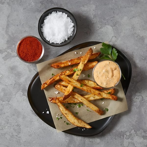 Seasoned fries with a side of creamy sauce, paprika, and sea salt on a black plate.