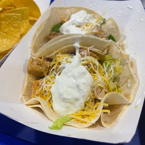 Two Yallah Taco chicken tacos with cheese, lettuce, and sour cream, with tortilla chips in the background.