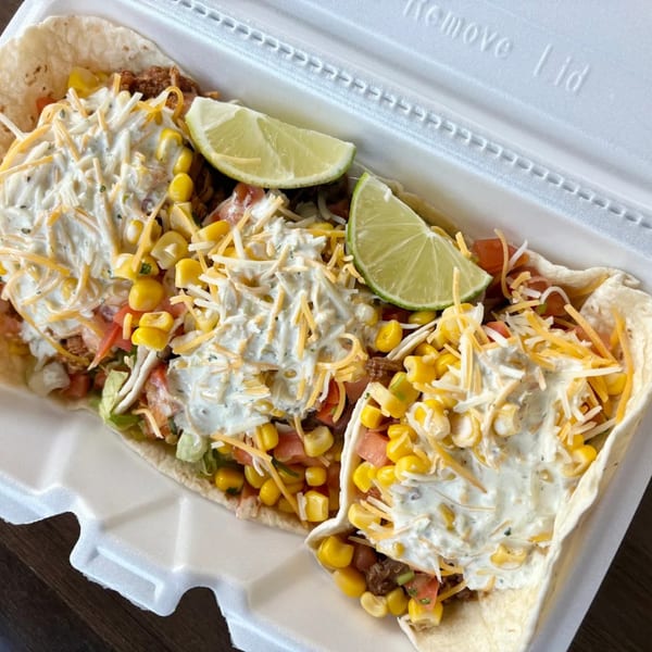 Two Yallah Taco soft tacos with meat, corn, cheese, and crema, garnished with lime wedges.