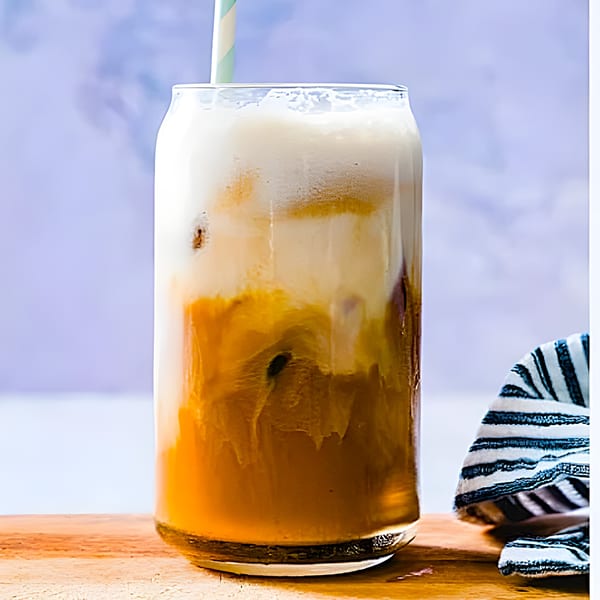 A tall glass of iced coffee with a foamy top and a striped straw on a wooden surface.