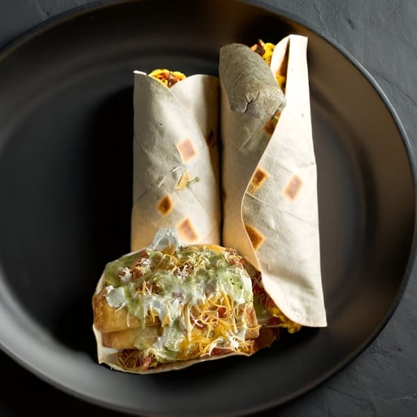 Two Yallah Taco burritos and a loaded burrito bowl with green and white sauce on a dark plate.